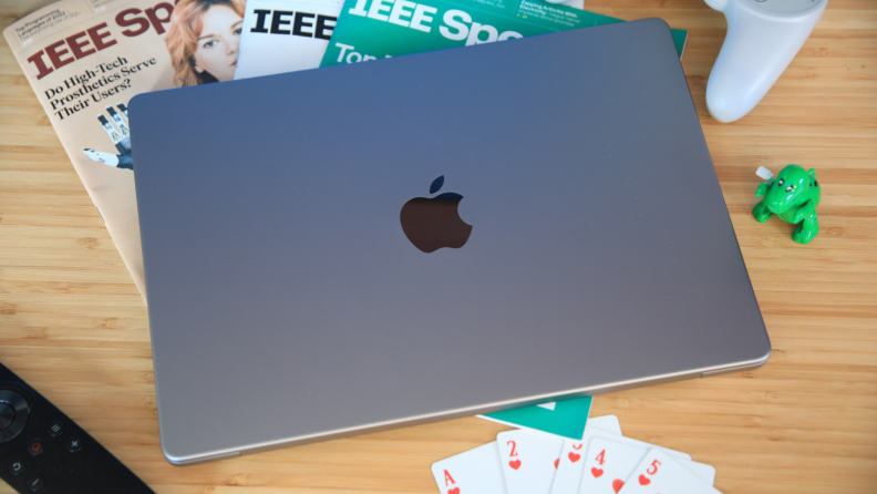 The Apple MacBook Pro 14 (2023) sitting on a wood table, closed, next to playing cards, magazines, and a green, toy dinosaur.